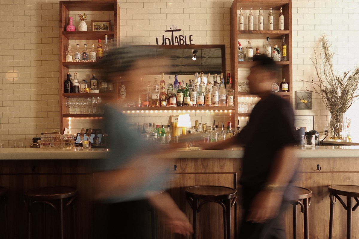 Bar area at Untable with bottle-lined shelves, subway tile, and bartenders in motion behind the counter
