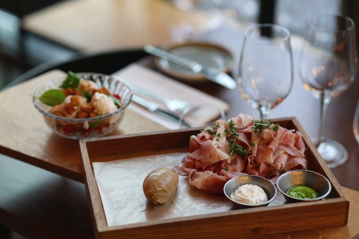 Wooden tray with sliced cured ham, dips, and a bowl of salad on a restaurant table with wine glasses