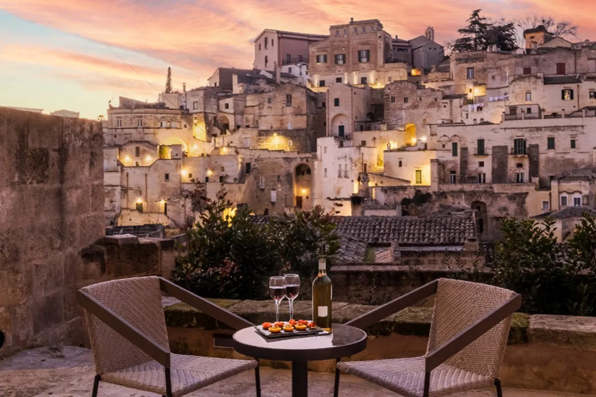 Terrace with table, chairs, wine and view over Matera's old town at dusk, hotel wall visible on the left