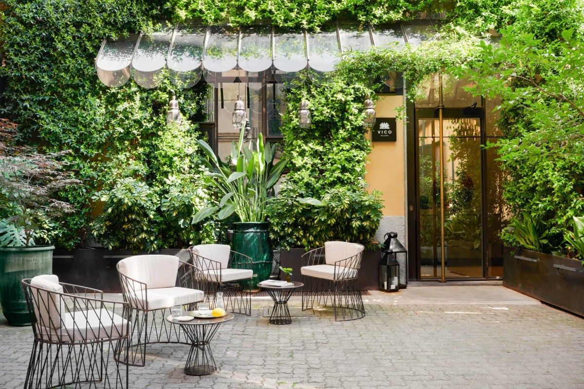 Daytime courtyard terrace with ivy-covered facade, outdoor seating, and Vico Milano sign by the entrance