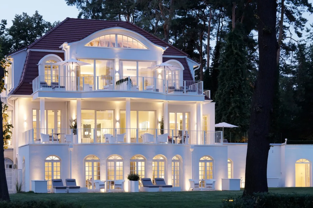 Illuminated white villa facade at dusk with arched windows, balconies and terrace seating, trees in background