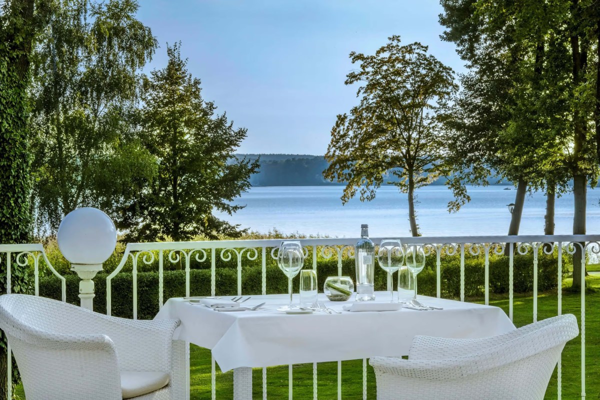 Outdoor dining table set for two on a terrace railing overlooking a lake surrounded by trees