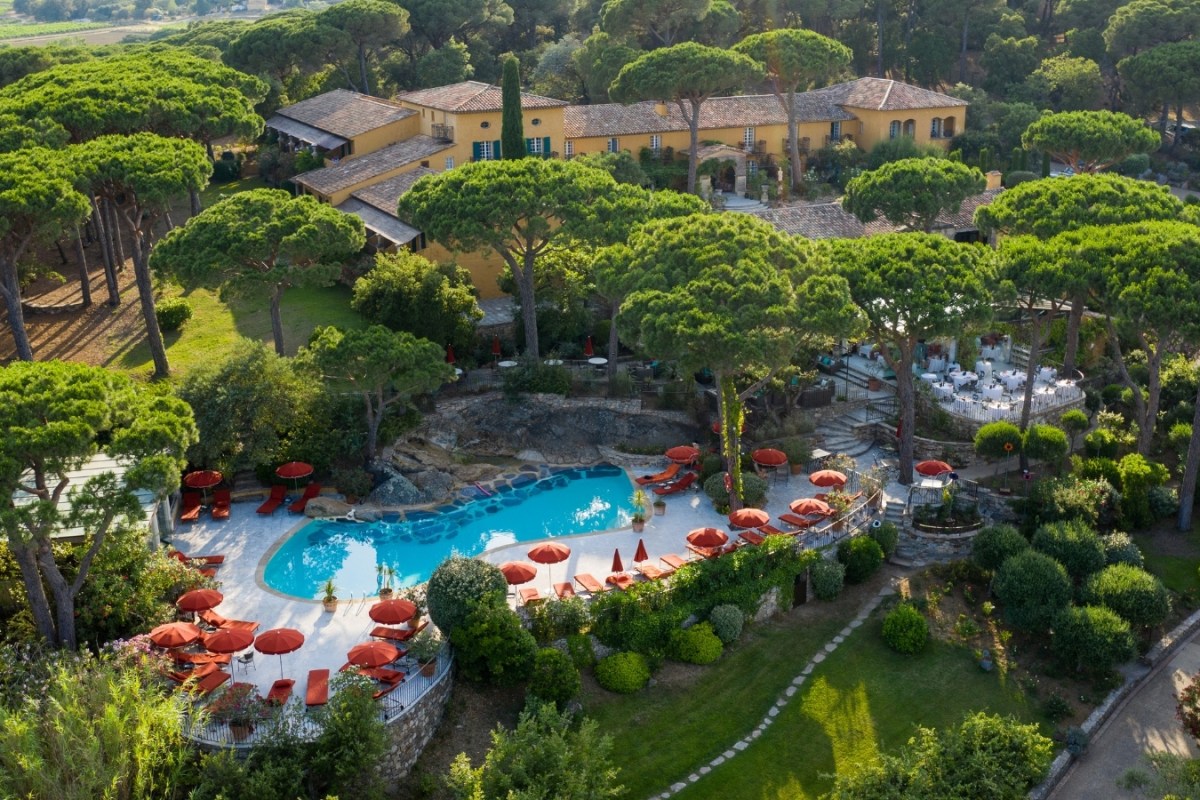 Aerial view of yellow villa hotel surrounded by pine trees with curved pool and red umbrellas