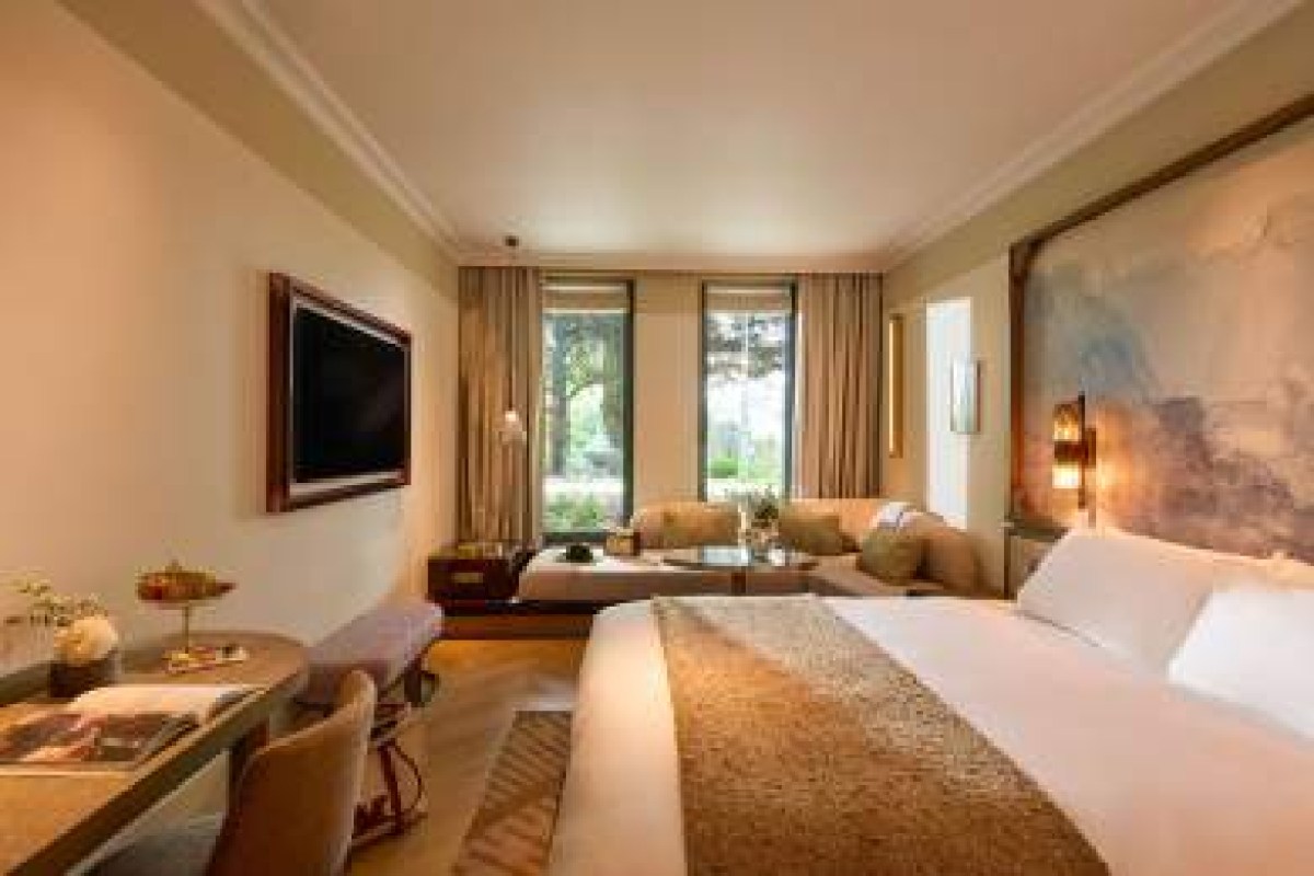 Spacious guest room with large bed, seating area, flatscreen TV, and tall windows overlooking greenery