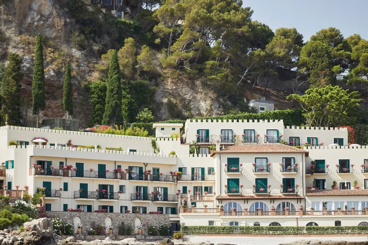 The cream façade of Villa Sant'Andrea with green shutters and crenellated rooftops, seen from the Bay of Mazzarò below wooded cliffs