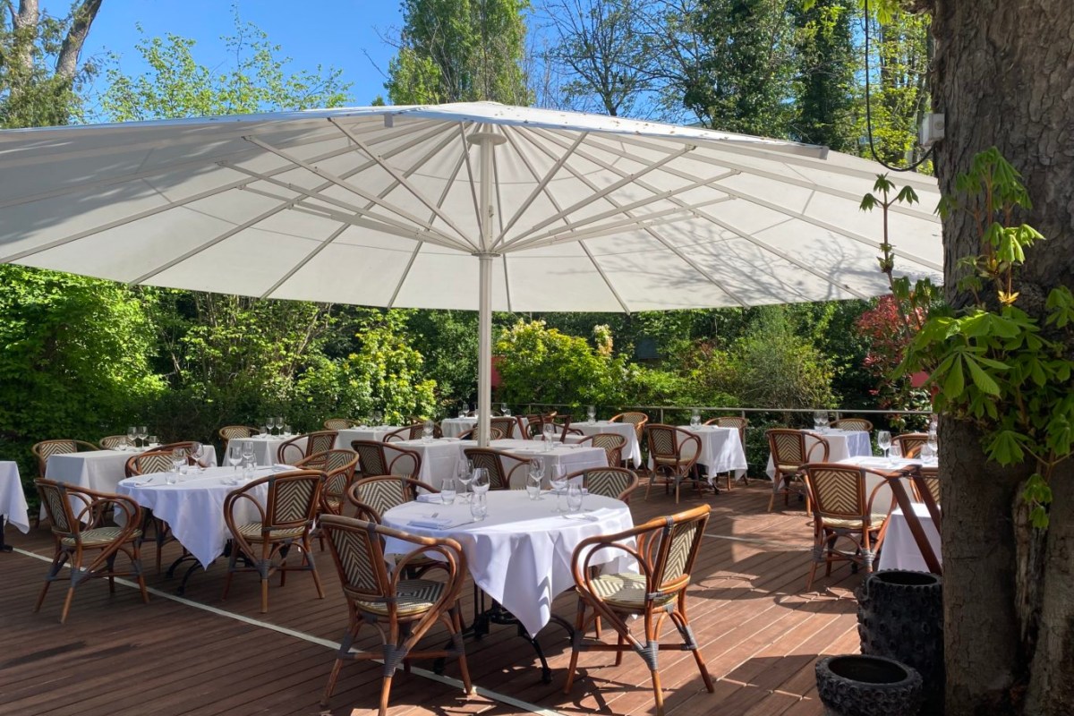 Outdoor terrace with white tablecloths, rattan chairs, and large umbrella amid greenery
