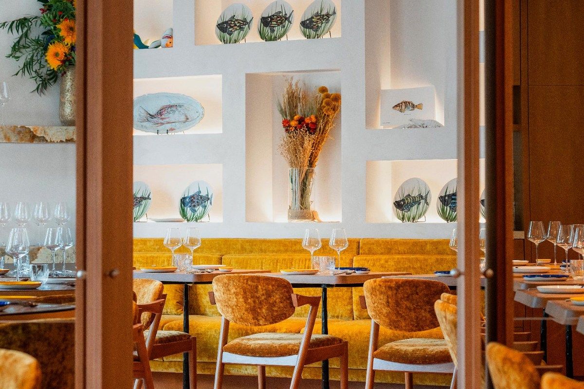 Dining room seen through a doorway, yellow velvet banquette and chairs, set tables, fish-themed ceramic plates in wall niches