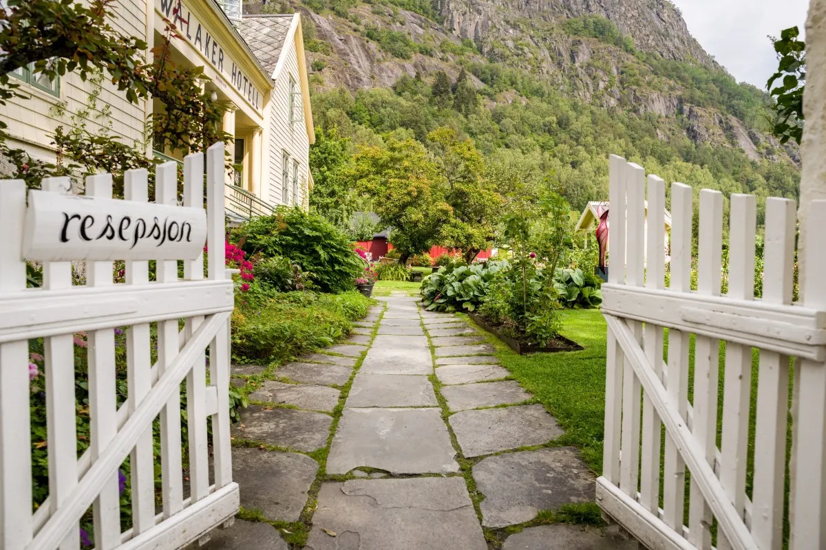 Garden path leading through open white gate to Walaker Hotell, with lush plants, lawn and mountain backdrop.