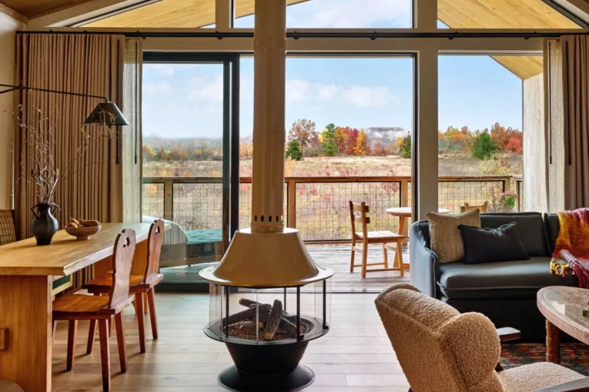 Guest suite living area with a central fireplace, dining table, and large windows showing fall foliage
