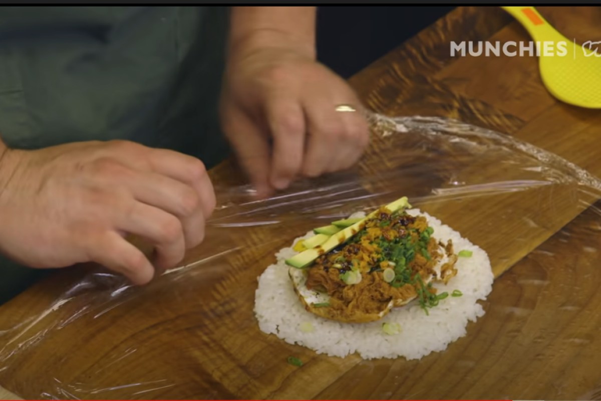 Close-up of hands assembling a rice dish with shredded meat, avocado, and scallions on a wooden board