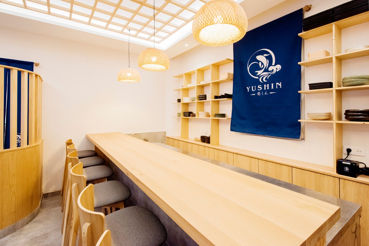Wooden counter seating area with hanging rattan lamps and a blue Yushin banner on the wall