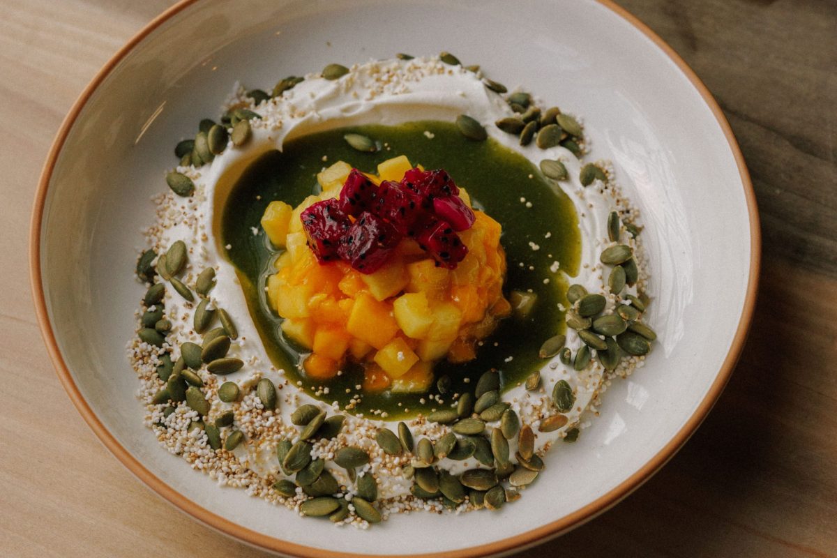 Overhead shot of a plated dessert with diced mango, dragon fruit, yogurt, and pumpkin seeds on a wooden table