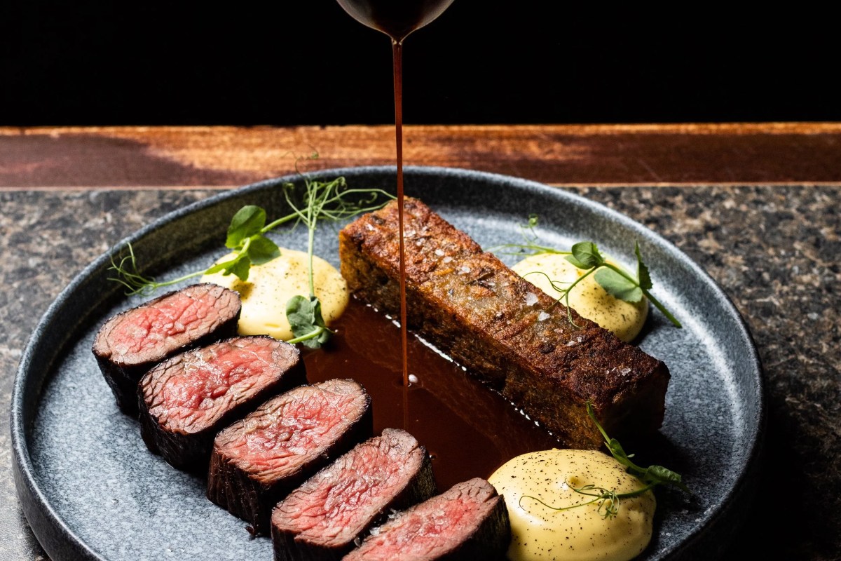 Sliced steak with sauce being poured, yellow purée dots, on a dark plate
