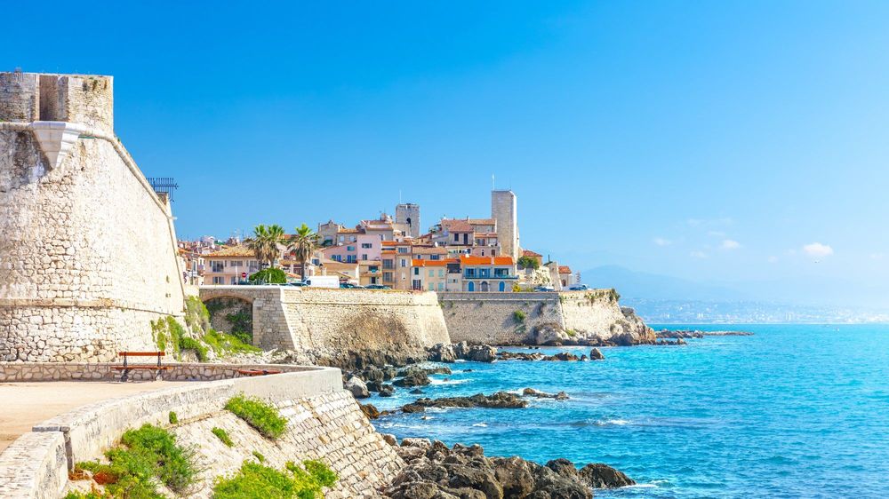 The sea walls of Antibes in bright daylight, the old town's towers and ochre houses above the rocks and the turquoise sea