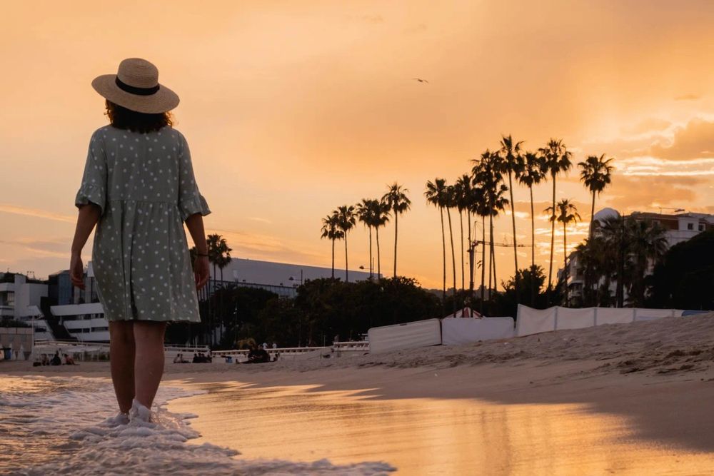 A walker in a sun hat on the sand at sunset in Cannes, palms silhouetted against an orange sky