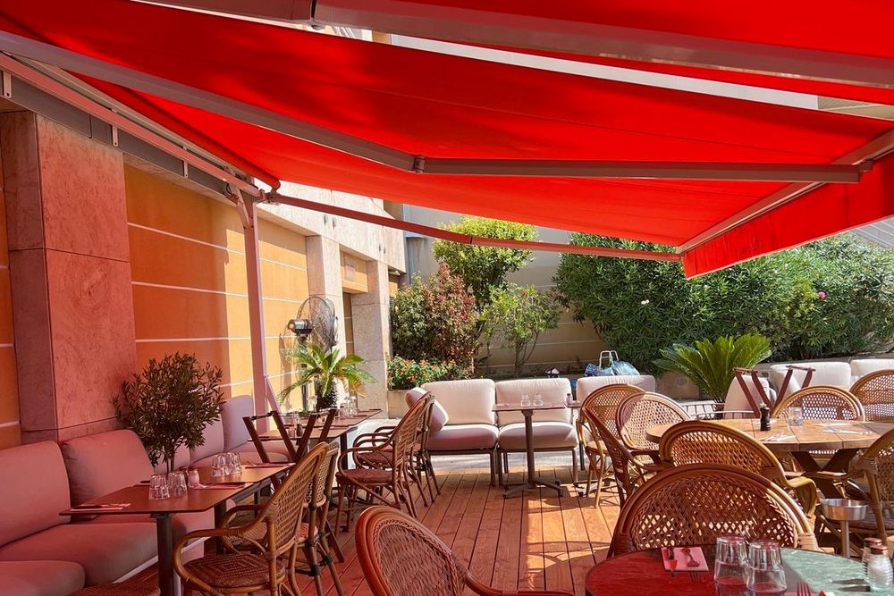 Cushioned benches and bistro chairs under the red awning of Chez Tonton's terrace, Cap-d'Ail