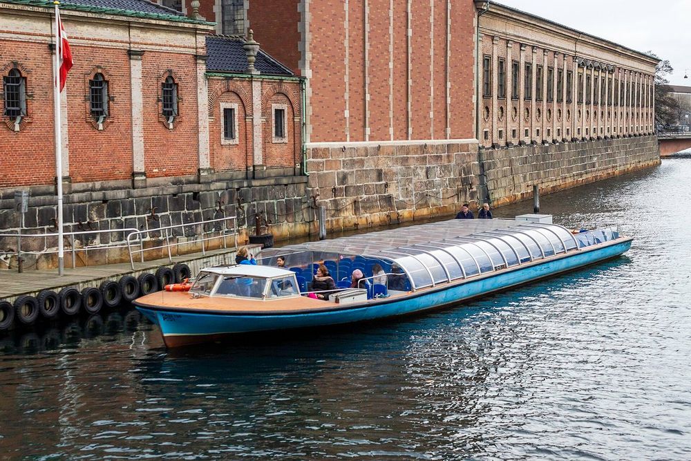 A blue-and-white Netto-Bådene canal boat moored in Børsgraven beside the brick walls of the old Stock Exchange