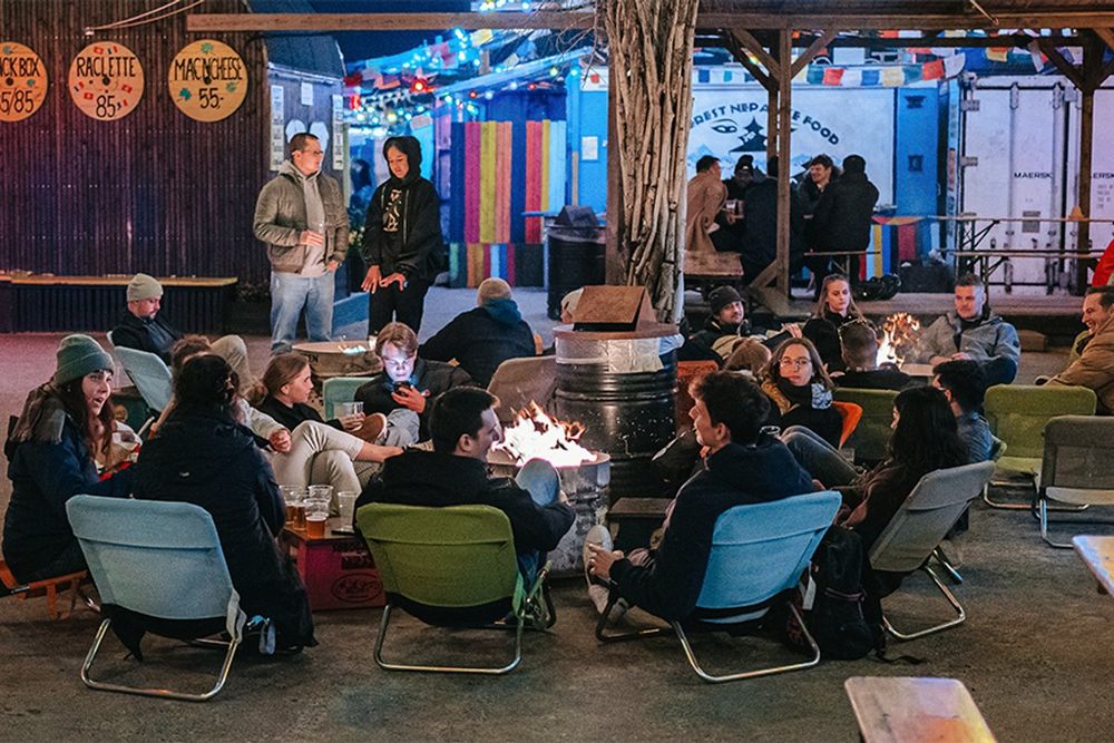 People in camping chairs around a fire pit among the colourful containers of Reffen street-food market in the evening