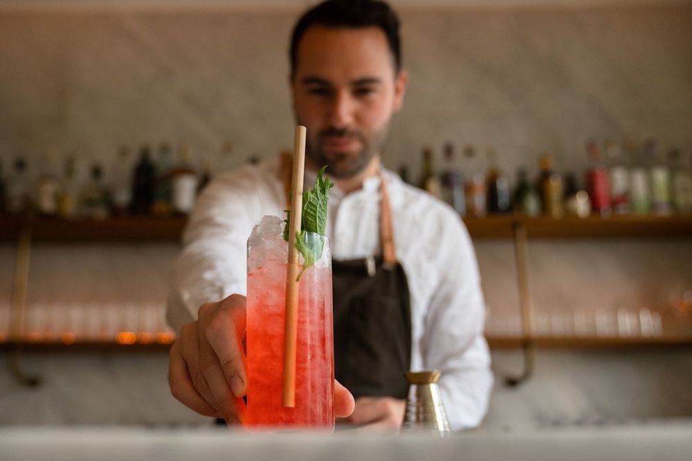 A bartender at Ruby holding out a pink cocktail garnished with rosemary