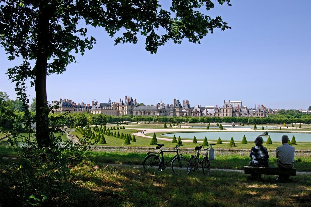 Visitors resting on a bench with bicycles under a tree, looking over the Grand Parterre to the château