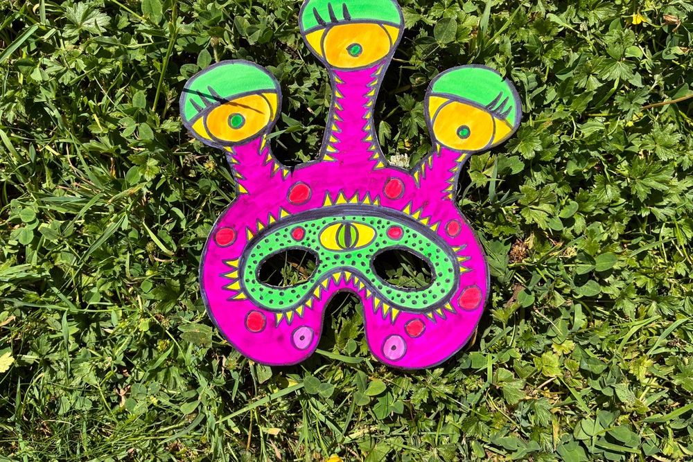 A bright pink three-eyed monster mask made in a children's workshop, laid on the grass