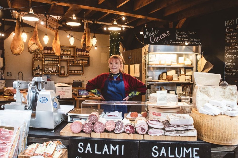 A trader behind a counter of salami, hams and cheeses at a stall in Borough Market