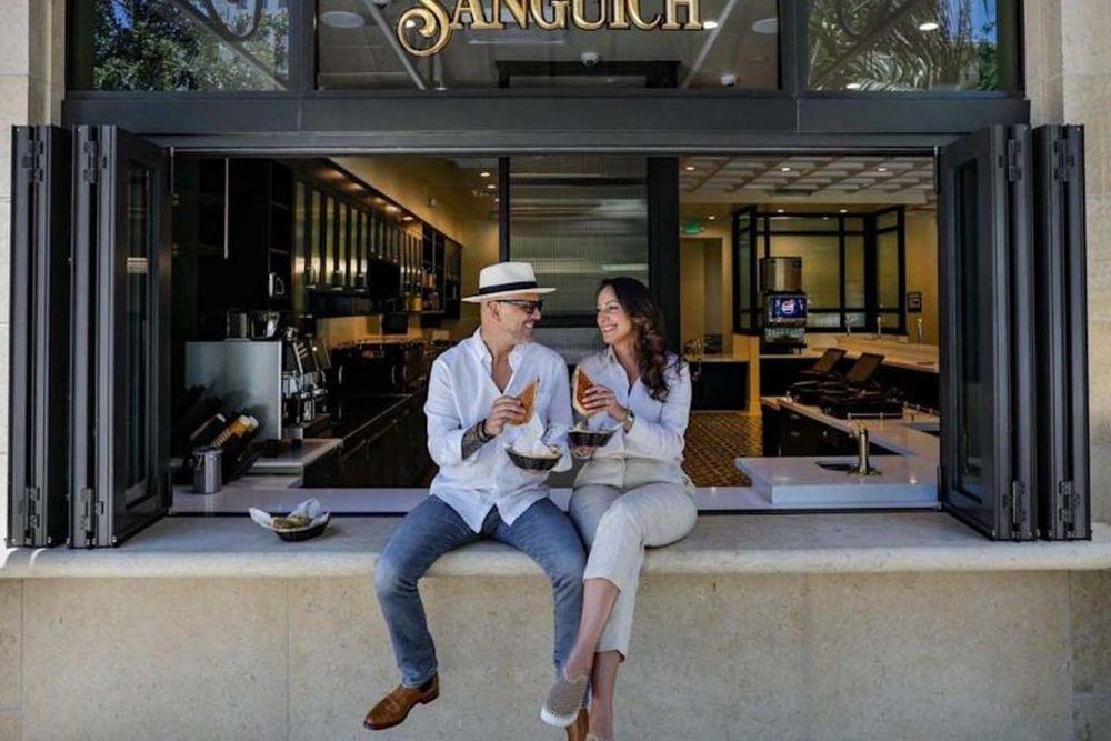 Two people eating Cuban sandwiches on the ledge of Sanguich's open walk-up window