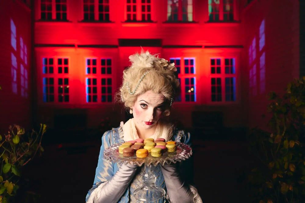 A performer in a powdered wig and 18th-century costume holding a tray of macarons before a red-lit house at Norsk Folkemuseum's Halloween