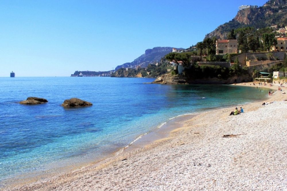 Pebbles and calm turquoise water at Plage du Golfe Bleu, with Roquebrune's wooded headland and Monaco beyond