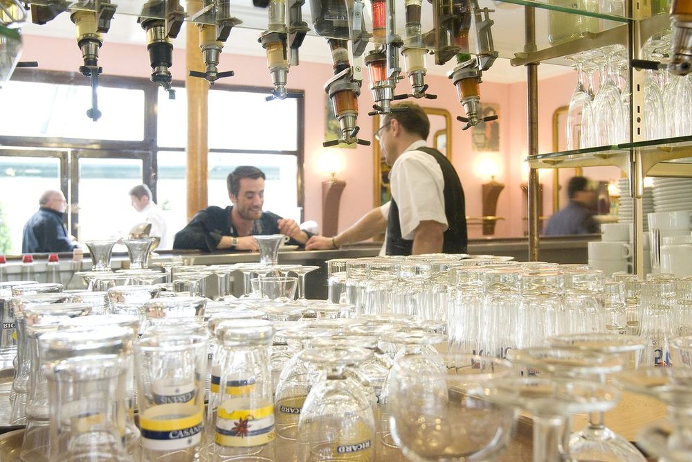 Rows of glasses on the bar of the Café de la Place, with a waiter and customers behind