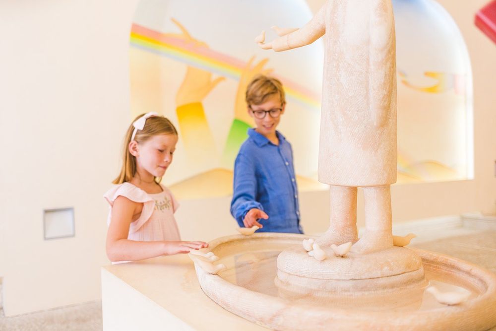Two children looking at a white Folon sculpture and font in the Chapelle des Pénitents Blancs