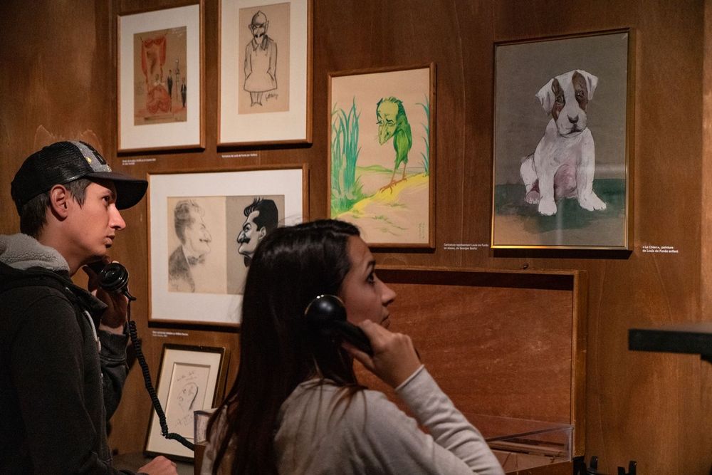 Visitors listening on old telephones beside drawings at the Musée Louis de Funès