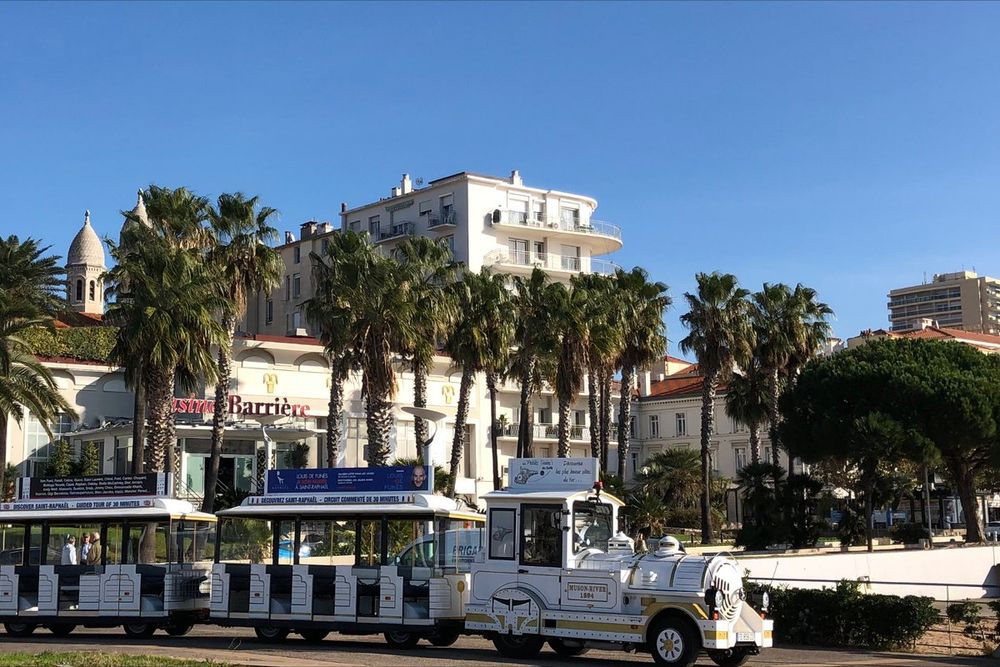 The white tourist train passing palms on the Saint-Raphaël seafront, with the basilica dome behind