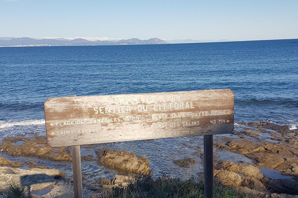 A weathered 'Sentier du Littoral' sign on the rocks with the gulf of Saint-Tropez behind