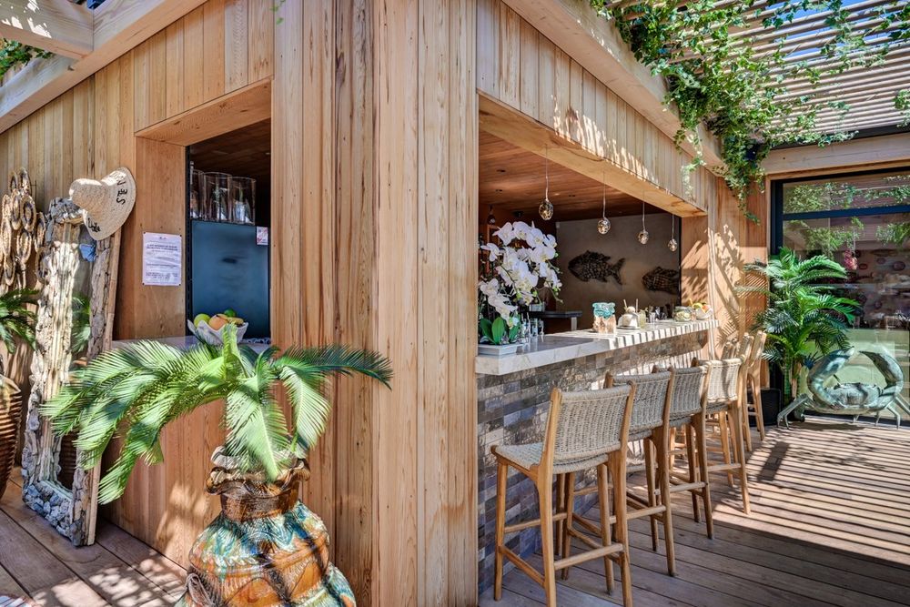 The wooden bar with high stools and tropical plants at La Voile beach club