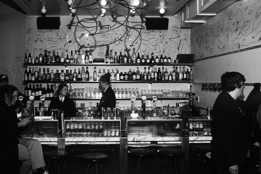 Bartenders and guests at the long bar of Bar Zeppelin on the top floor of Gondolen, in black and white