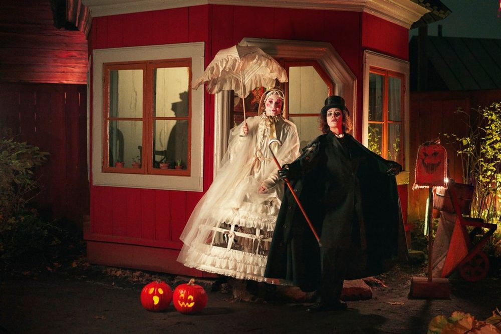 Two ghostly Halloween characters in Victorian dress outside a red wooden house at Skansen, lit by jack-o'-lanterns