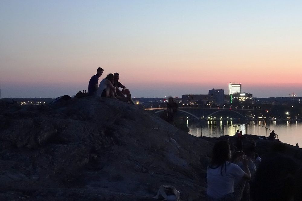 People sitting on the rocks of Skinnarviksberget against a pink and orange sky after sunset, the city lights beyond