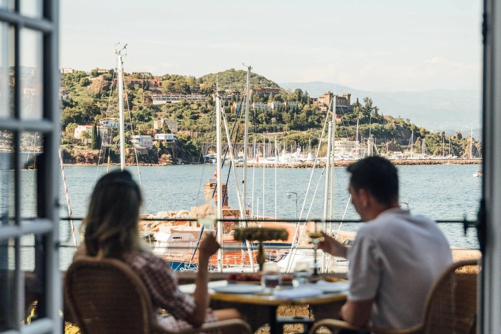 Two guests at a table on the Château de Théoule terrace, looking out over the port's masts to the green Estérel hills
