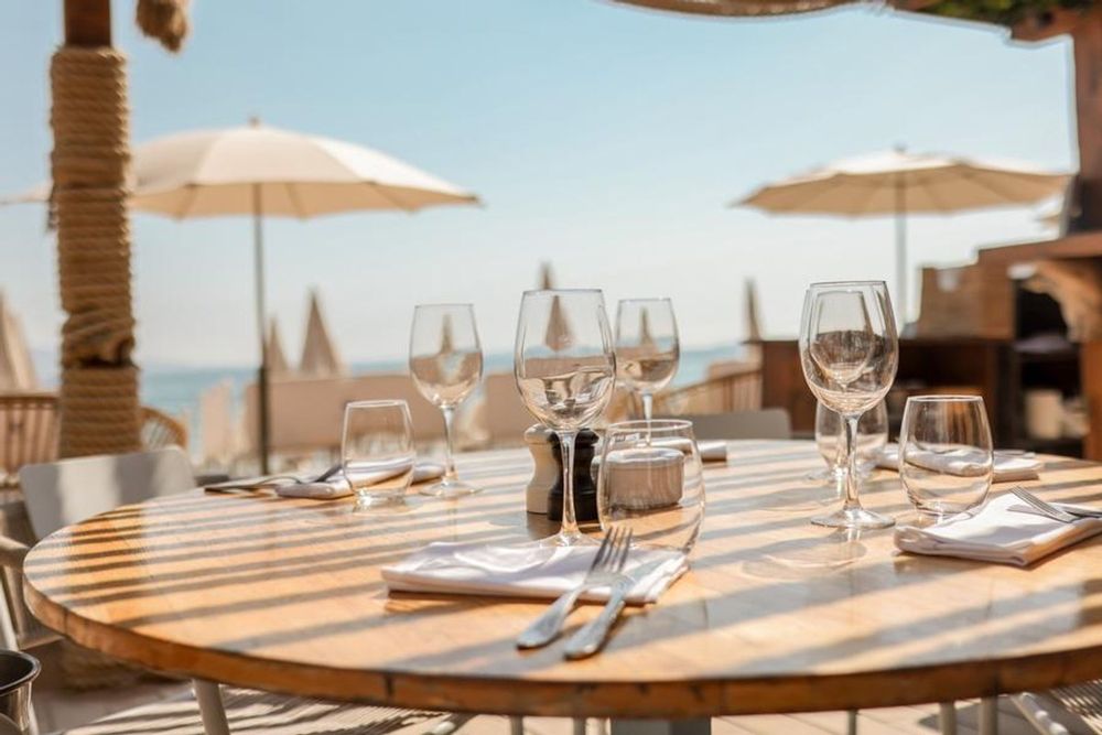 A laid round table under parasols on the Marco Polo terrace, with the sea and the bay beyond