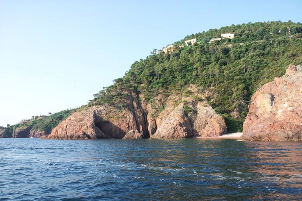 Red cliffs and wooded slopes of the Pointe de l'Aiguille seen from the sea, with a small beach at their foot