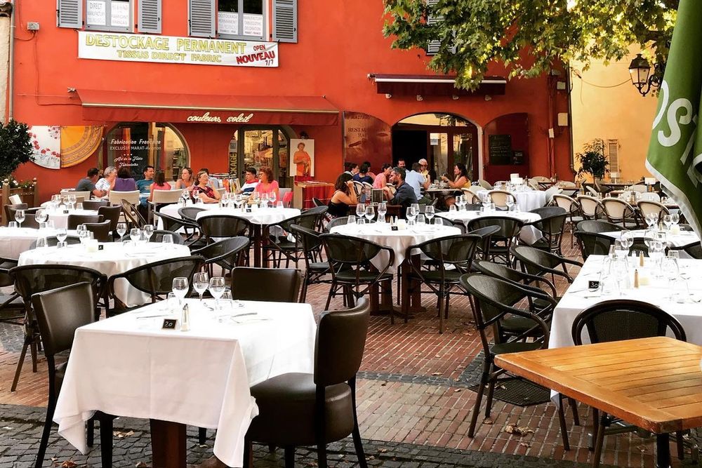 White-clothed tables on the square terrace of Le Café de la Poste in front of its red façade in Biot