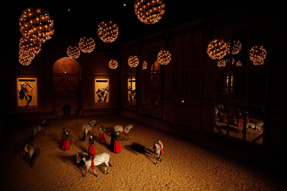 Riders in red under hanging lanterns in the riding hall of the Académie équestre de Versailles
